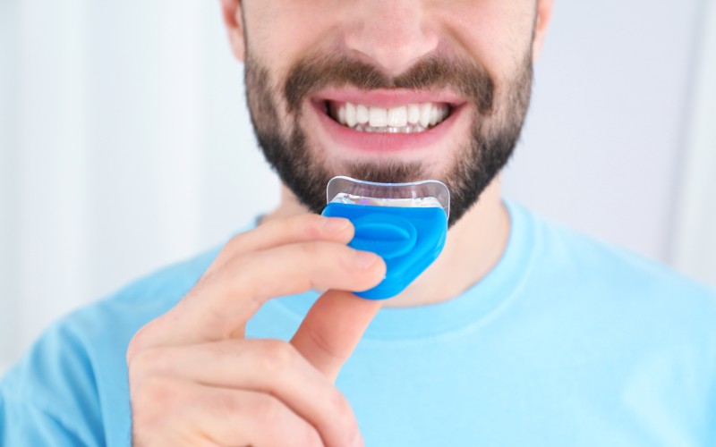 young man using teeth-whitening device on light background closeup